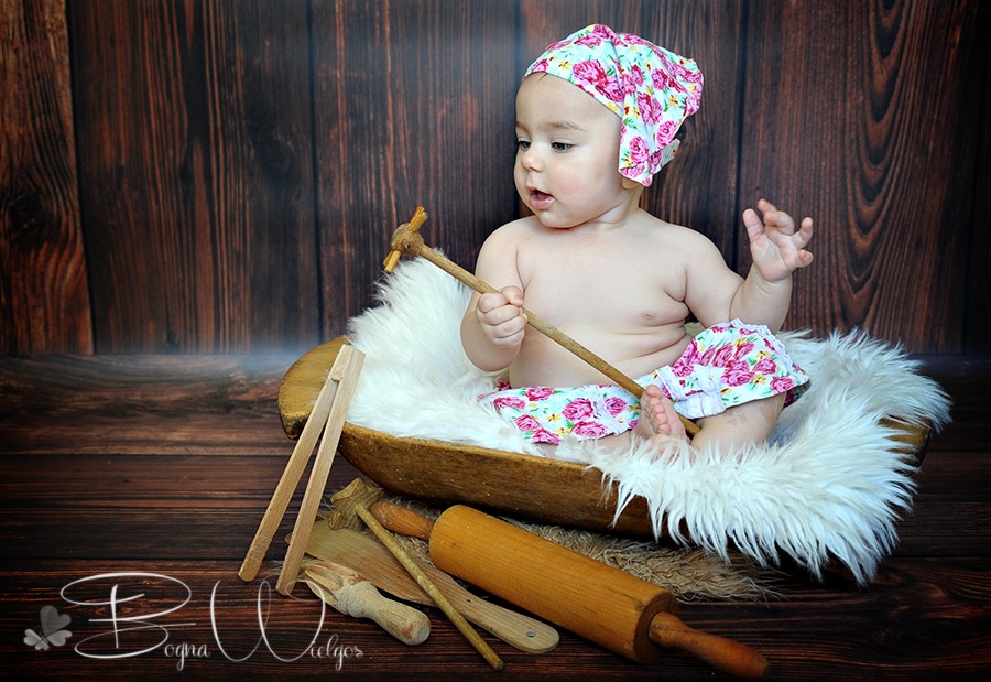 Niemowlę w kwiecistej czapeczce i majteczkach siedzi w drewnianej misce wyłożonej białym futerkiem, trzymając w rączce drewnianą zabawkę na tle drewnianej ściany i rekwizytów kuchennych.