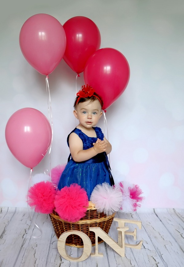 Dziewczynka w niebieskiej sukience i czerwonej opasce siedzi w koszyku otoczonym różowymi pomponami i balonami, z drewnianym napisem 'ONE' z przodu.