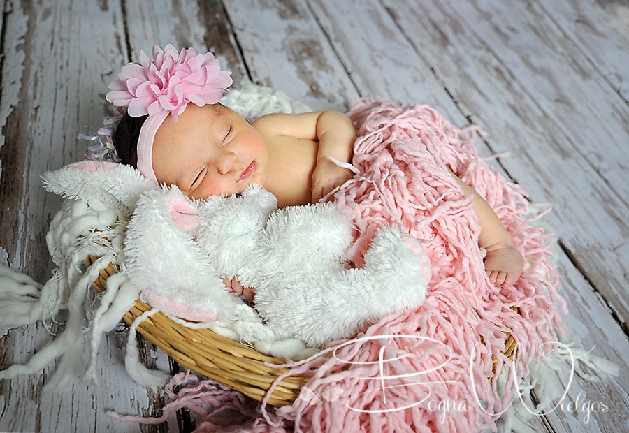 Śpiąca noworodka z różowym kwiatem na głowie, otulona w koszyku różową i białą włóczką, obok pluszowy królik z różowymi uszami, na tle drewnianej podłogi.
