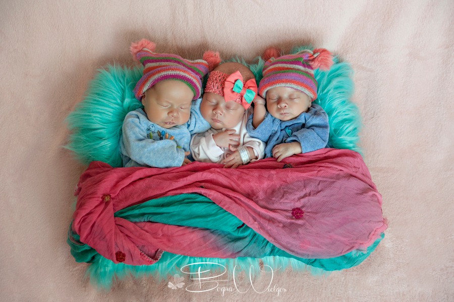 Troje śpiących noworodków, dwaj chłopcy w pasiaste czapki z pomponami i niebieskich ubrankach, dziewczynka z kokardą na głowie, owinięci kolorową tkaniną na turkusowym futerku.