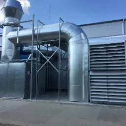 Zewnętrzna instalacja wentylacyjna z metalowymi rurami i kratkami wentylacyjnymi, widoczna na tle szarej ściany budynku i błękitnego nieba.