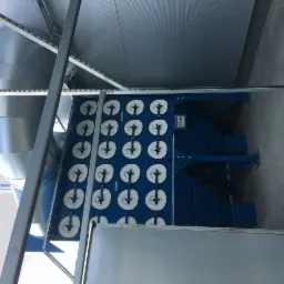 Niebieska centrala wentylacyjna z rzędem filtrów w białych obudowach, widoczny srebrny kanał wentylacyjny i konstrukcja wsporcza pod sufitem.