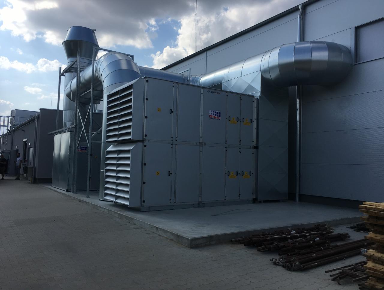 Instalacja wentylacji mechanicznej z rekuperacją na dachu budynku komercyjnego, widoczne kanały wentylacyjne i moduły centrali wentylacyjnej, częściowo widoczne elementy montażowe na pierwszym planie.