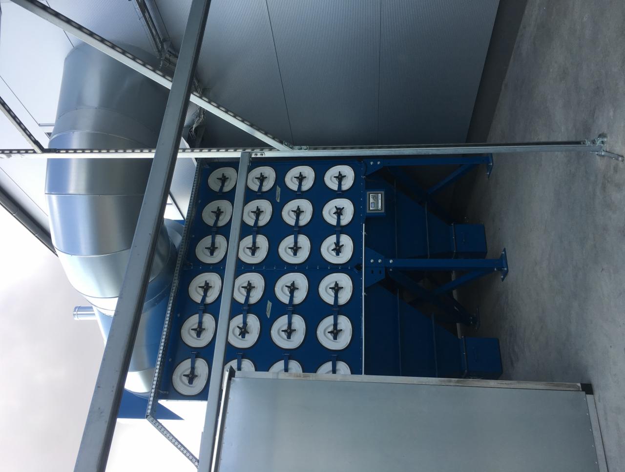 Niebieska centrala wentylacyjna z rzędem filtrów w białych obudowach, widoczny srebrny kanał wentylacyjny i konstrukcja wsporcza pod sufitem.