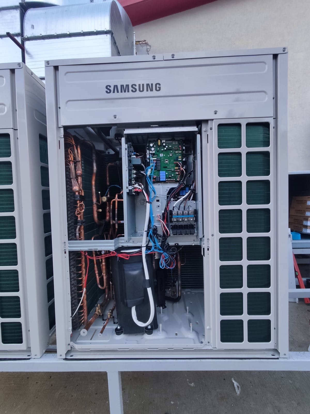 Otwarta obudowa klimatyzacji Samsung VRF ukazuje wewnętrzne komponenty: miedziane rurki, moduł sterujący z elektroniką i okablowaniem. Widoczne elementy montażowe i podłączeniowe.