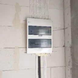 Biała skrzynka rozdzielcza z podłączonymi przewodami na ścianie z betonu komórkowego, uszczelniona pianką montażową.