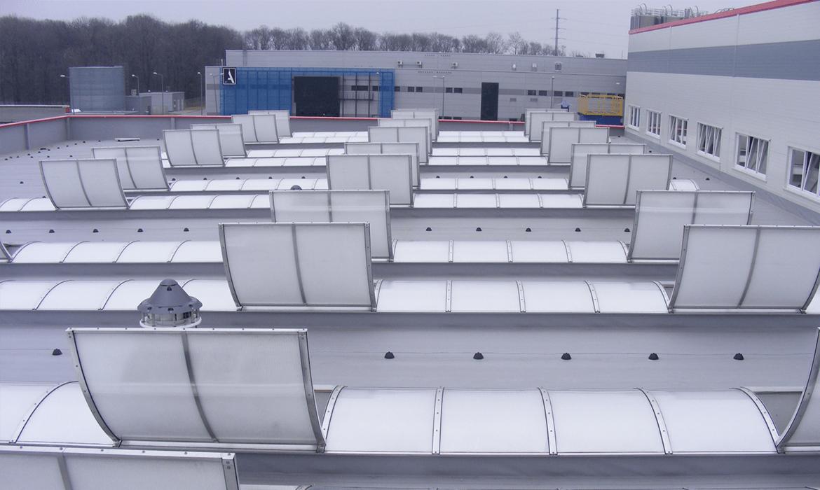 Widok z góry na płaski dach przemysłowy z licznymi świetlikami dachowymi, wentylacją i elementami konstrukcyjnymi, na tle budynków przemysłowych i drzew bez liści pod pochmurnym niebem.