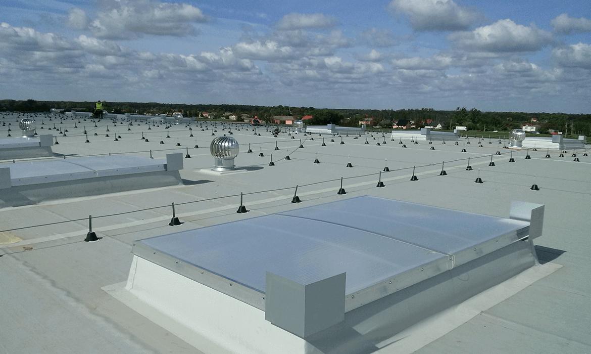 Widok z góry na płaski dach przemysłowy z szarym pokryciem, świetlikami dachowymi, turbinami wiatrowymi i systemem zabezpieczeń linowych z czarnymi punktami kotwicznymi. W tle widoczna zieleń...