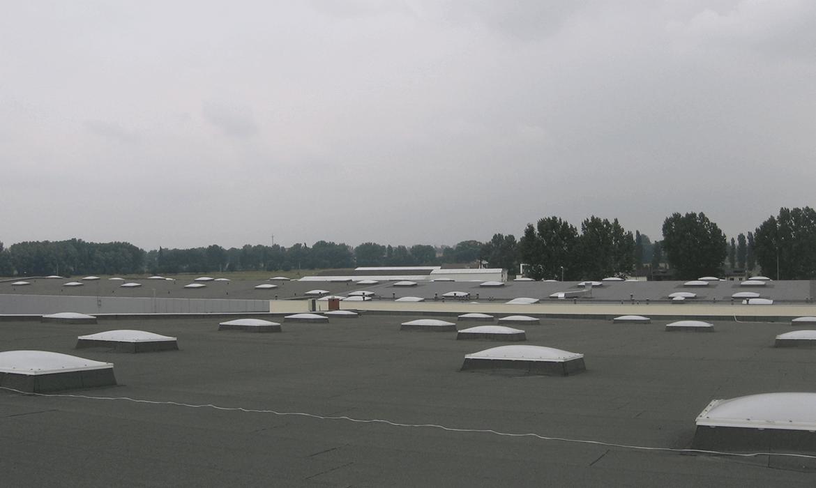 Rozległy, płaski dach przemysłowy pokryty papą z licznymi świetlikami kopułowymi, widok z góry w pochmurny dzień.