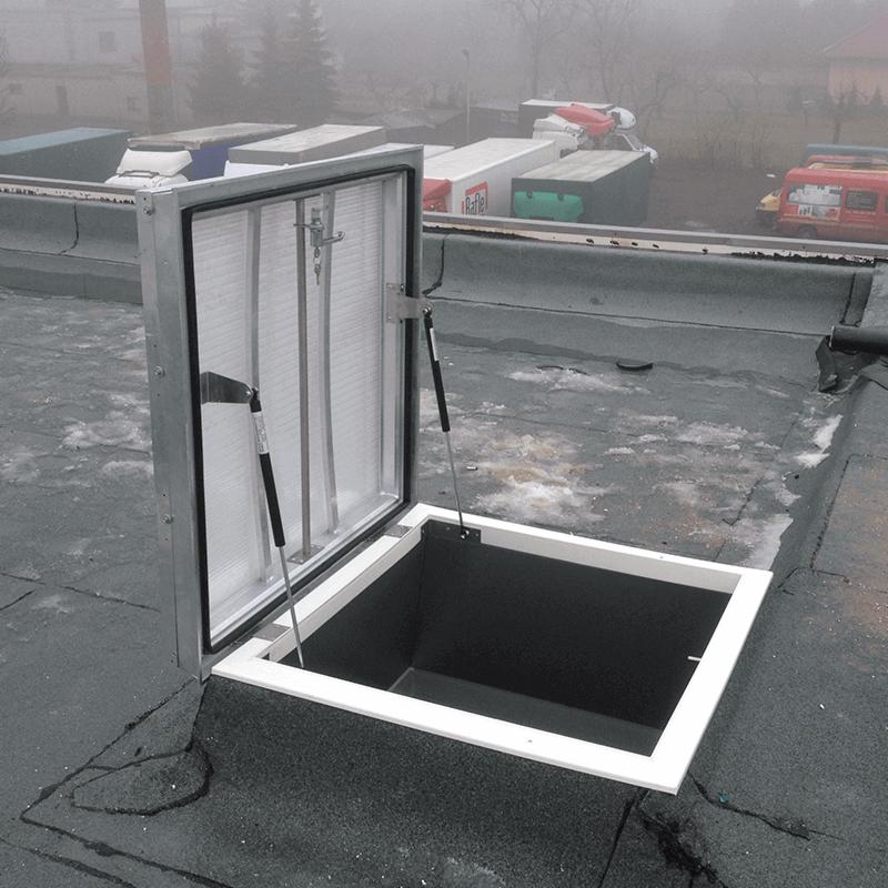 Otwarty, metalowy właz dachowy z widocznymi mechanizmami podtrzymującymi pokrywę na płaskim dachu pokrytym papą, z rozmytym tłem zabudowań i pojazdów w mglisty dzień.