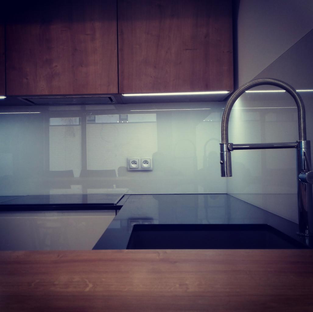 Nowoczesna kuchnia z drewnianymi szafkami wiszącymi, szklanym panelem nad blatem, chromowaną baterią i czarnym zlewem, oświetlona listwą LED pod szafkami.