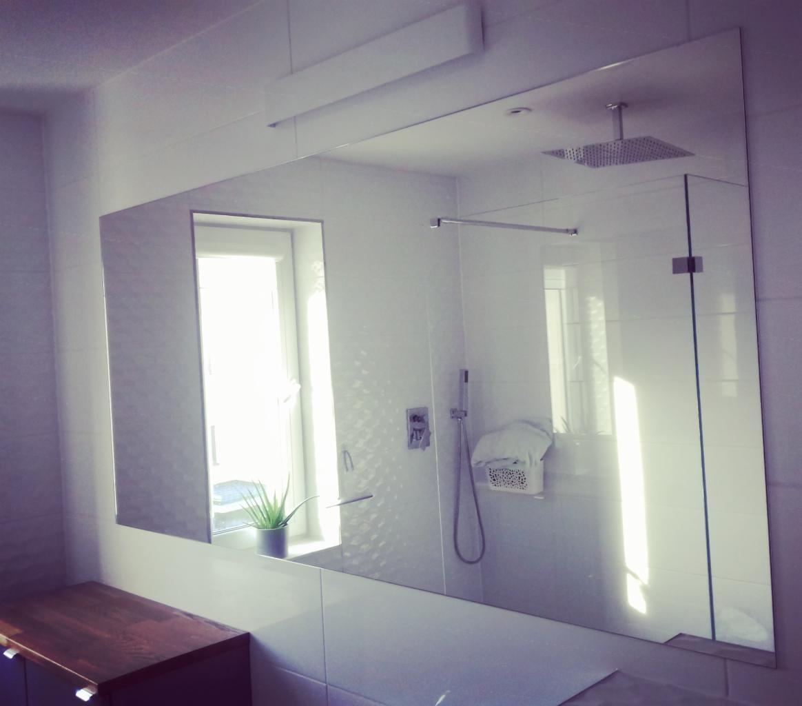 Duże lustro w nowoczesnej łazience z białymi płytkami, odbijające kabinę prysznicową typu walk-in z deszczownicą i okno z rośliną doniczkową na parapecie, oświetlone górnym światłem.