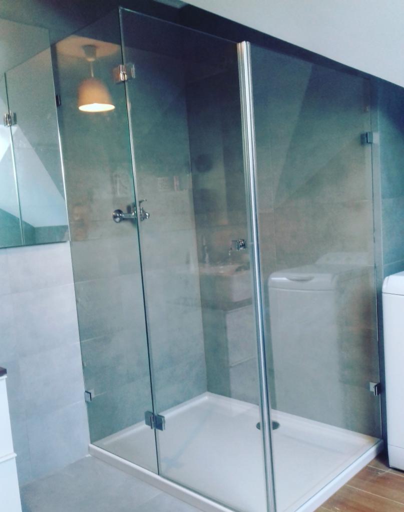 Szklana kabina prysznicowa z brodzikiem w szaro-białej łazience, widoczna pralka w tle, chromowane zawiasy i uchwyt, oświetlenie sufitowe włączone.