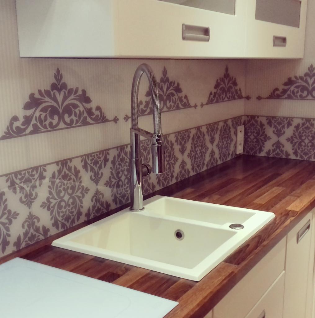 Biały, dwukomorowy zlew podwieszany z chromowaną baterią w kuchni w Drohiczynie. Nad zlewem dekoracyjne, szare wzory na ścianie, a blat z drewna klejonego.