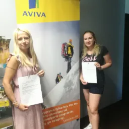 Dwie kobiety trzymające dokumenty na tle roll-upu reklamowego firmy ubezpieczeniowej Aviva, prezentującego wspinaczkę górską.