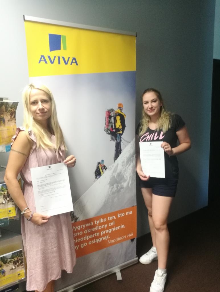 Dwie kobiety trzymające dokumenty na tle roll-upu reklamowego firmy ubezpieczeniowej Aviva, prezentującego wspinaczkę górską.