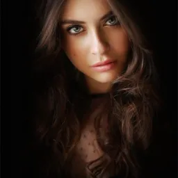 Portret kobiety z długimi, falowanymi włosami w ciemnym otoczeniu. Uwagę przykuwają jej duże, zielone oczy i delikatny makijaż. Subtelne oświetlenie modeluje rysy twarzy.