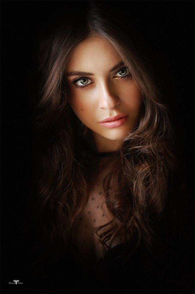 Portret kobiety z długimi, falowanymi włosami w ciemnym otoczeniu. Uwagę przykuwają jej duże, zielone oczy i delikatny makijaż. Subtelne oświetlenie modeluje rysy twarzy.