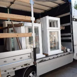 Biały samochód dostawczy z otwartą przestrzenią ładunkową, wypełnioną nowymi, białymi oknami zabezpieczonymi drewnianymi belkami podczas transportu.