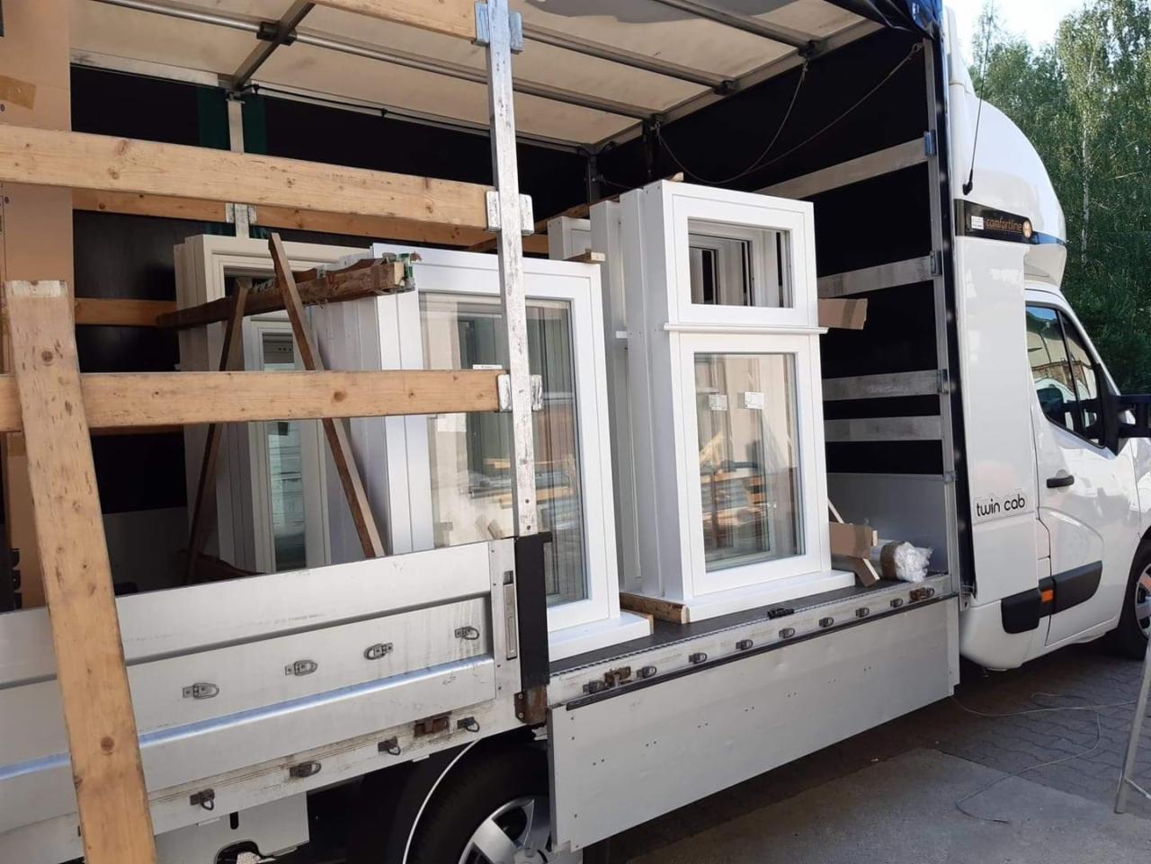 Biały samochód dostawczy z otwartą przestrzenią ładunkową, wypełnioną nowymi, białymi oknami zabezpieczonymi drewnianymi belkami podczas transportu.