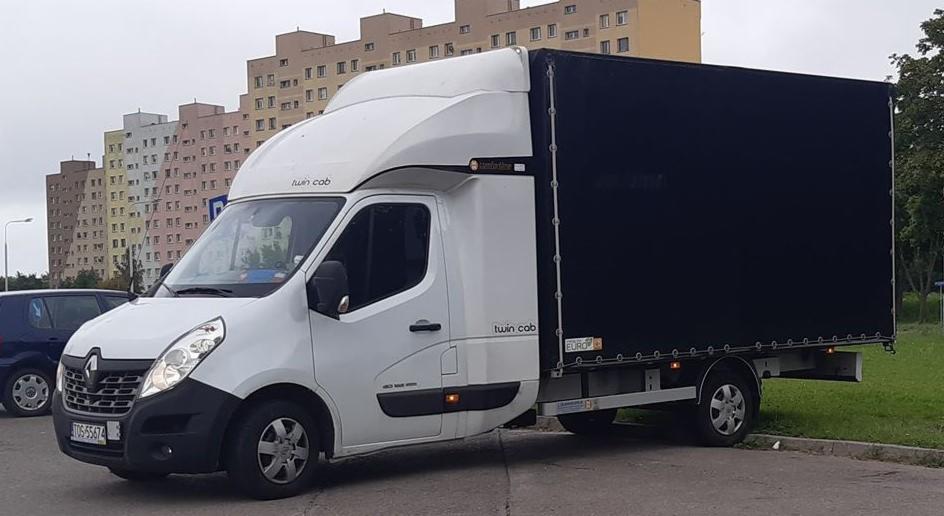 Biały samochód dostawczy Renault Master Twin Cab z czarną plandeką na tle bloku mieszkalnego, widoczny fragment tablicy rejestracyjnej.