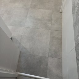 Art-Bud Remonty i Wykończenia - Jasnoszara, kwadratowa podłoga z płytek w łazience, widok z góry, z fragmentem ściany z dekoracyjnym wzorem i białej listwy przypodłogowej.