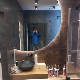 Art-Bud Remonty i Wykończenia - Nowoczesna łazienka z umywalką nablatową, podświetlanym lustrem o nieregularnym kształcie i kabiną prysznicową, widoczne tuby silikonu na blacie, odbicie osoby robiącej zdjęcie w lustrze.