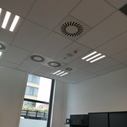 Art-Bud Remonty i Wykończenia - Biały sufit podwieszany z rastrowymi lampami LED i okrągłymi kratkami wentylacyjnymi w biurze, widok z dołu, fragment okna i szafki w tle.