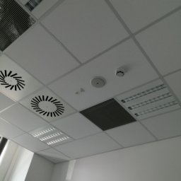 Art-Bud Remonty i Wykończenia - Sufit podwieszany z panelami, kratkami wentylacyjnymi, czujnikiem dymu i oświetleniem rastrowym w biurze, widziany pod kątem.