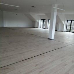 Art-Bud Remonty i Wykończenia - Przestronne, jasne wnętrze biurowe z panelami podłogowymi imitującymi drewno, białymi ścianami i kolumnami oraz dużymi oknami z czarnymi ramami.