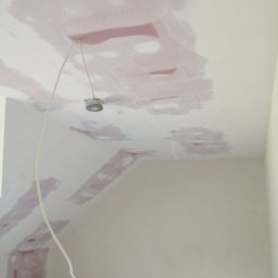 Art-Bud Remonty i Wykończenia - Sufit w trakcie prac wykończeniowych z widocznymi łatami siatki wzmacniającej i nałożoną warstwą szpachli, przygotowany pod malowanie, z wystającym przewodem elektrycznym.