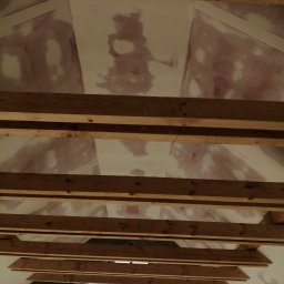 Art-Bud Remonty i Wykończenia - Montaż sufitu podwieszanego z drewnianą konstrukcją nośną, widoczne łączenia płyt szpachlowane, w tle okno z widokiem na zieleń.