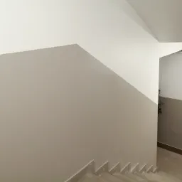 Klatka schodowa po malowaniu, ściany dwukolorowe: kremowa g&oacute;ra i beżowy d&oacute;ł, widoczne schody i fragment drewnianej balustrady.