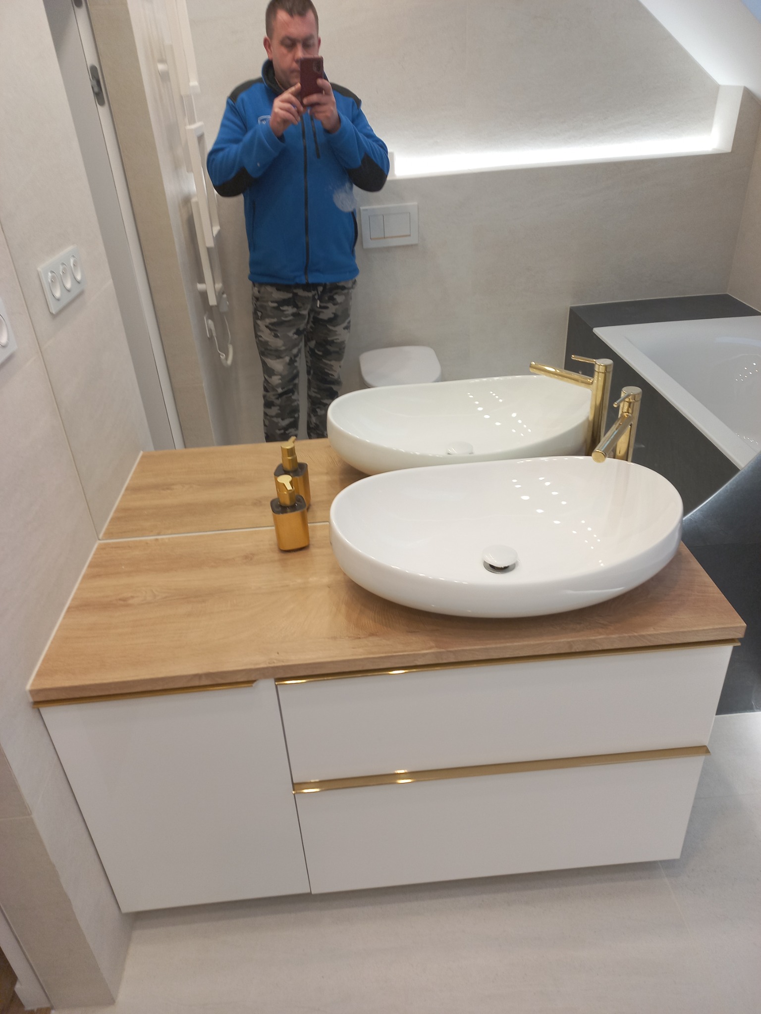 Nowoczesna łazienka z podwójną umywalką nablatową, drewnianym blatem i złotymi bateriami, odbicie fotografa w lustrze.