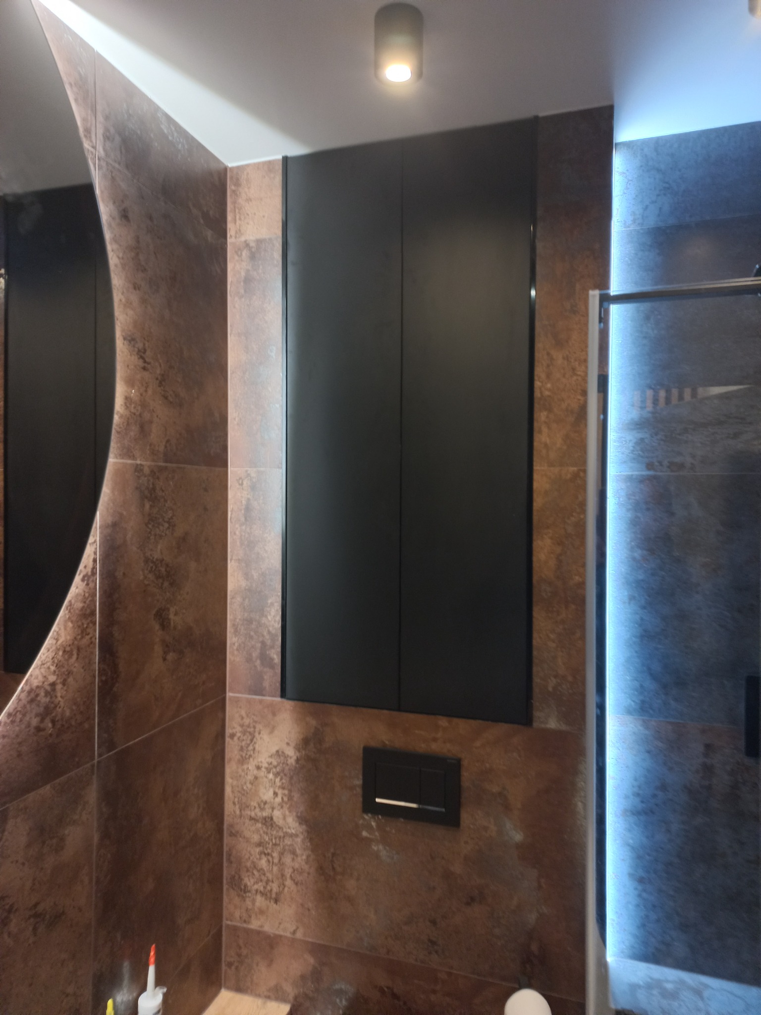 Nowoczesna łazienka z brązowymi płytkami imitującymi rdzę, czarnymi szafkami wiszącymi i podświetlaną kabiną prysznicową.