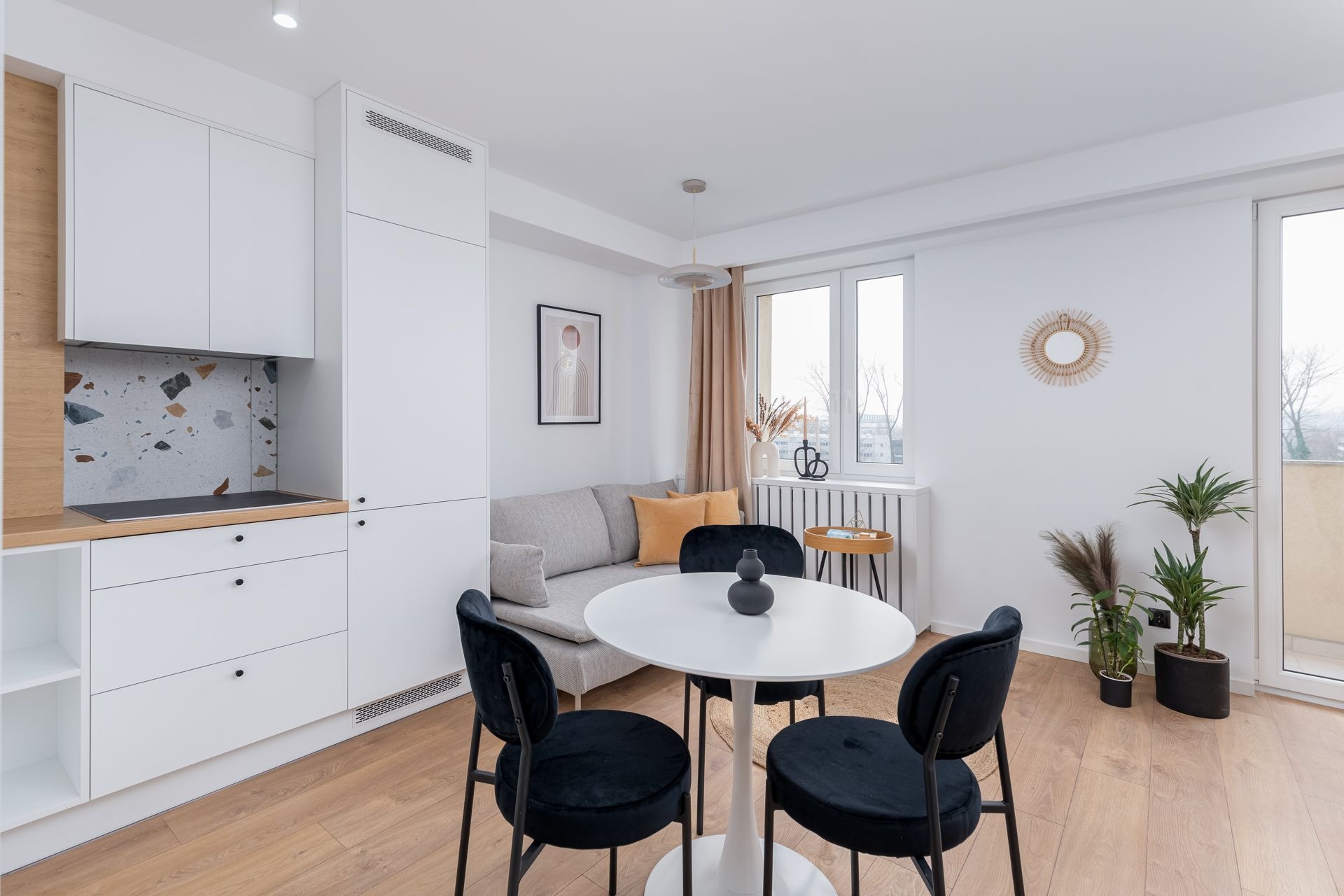 Jasne, nowocześnie urządzone wnętrze mieszkania z aneksem kuchennym, okrągłym stołem z czarnymi krzesłami i szarą sofą z pomarańczowymi poduszkami.