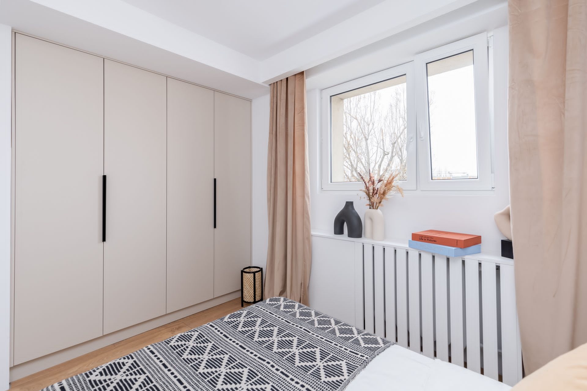 Jasna sypialnia z beżową szafą wnękową o minimalistycznych, długich, czarnych uchwytach, okno z beżowymi zasłonami, dekoracyjny grzejnik z książkami i wazonami, wzorzysta narzuta na łóżku.