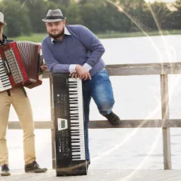 Dwaj mężczyźni w kapeluszach prezentują instrumenty muzyczne: akordeon i keyboard, stojąc na drewnianym pomoście nad wodą, z efektem świetlnym imitującym fajerwerki.