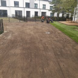 Proxential sp. z o.o. - Prace ziemne przed domem, przygotowanie podłoża i układanie pierwszego pasa trawy z rolki przez trzech pracowników w odzieży roboczej, widoczny fragment drewnianego tarasu.