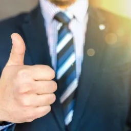 Mężczyzna w garniturze z krawatem pokazuje kciuk w górę, symbolizując aprobatę lub pozytywną opinię.