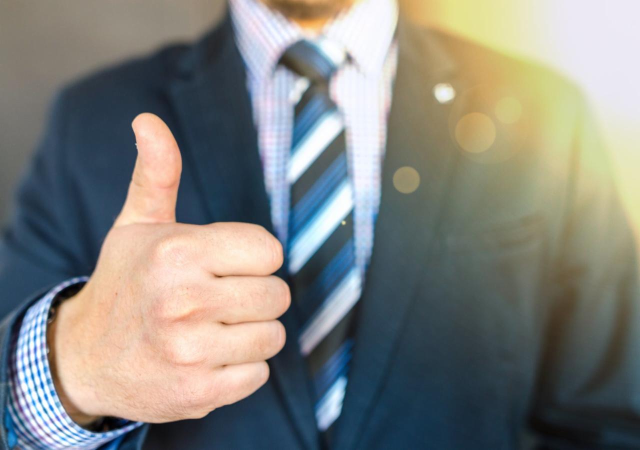 Mężczyzna w garniturze z krawatem pokazuje kciuk w górę, symbolizując aprobatę lub pozytywną opinię.