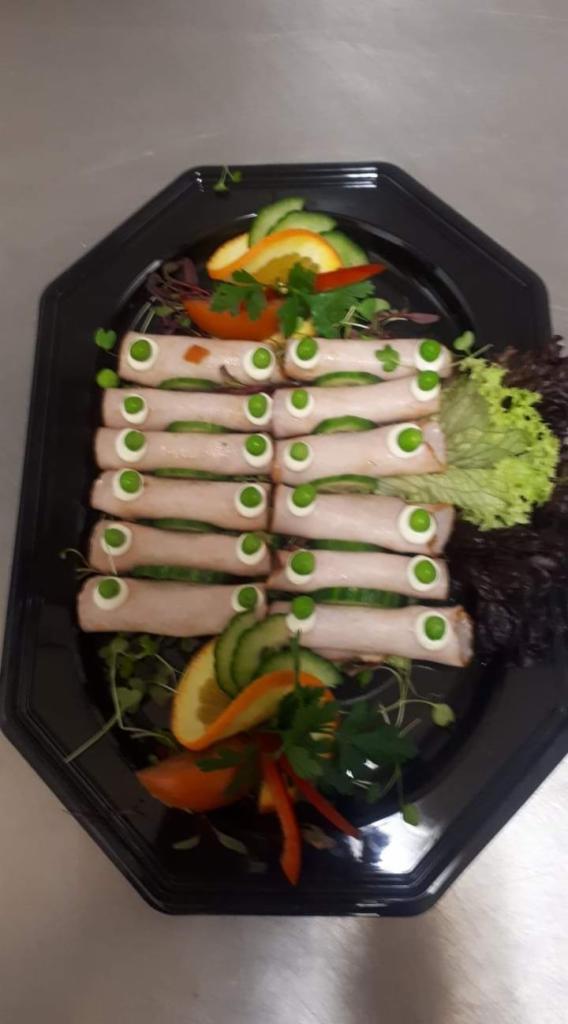 Dekoracyjnie ułożone roladki z szynki z plasterkiem ogórka i kleksem majonezu z groszkiem na wierzchu, ułożone na czarnym talerzu z dodatkiem pomarańczy, ogórka, papryki i sałaty.