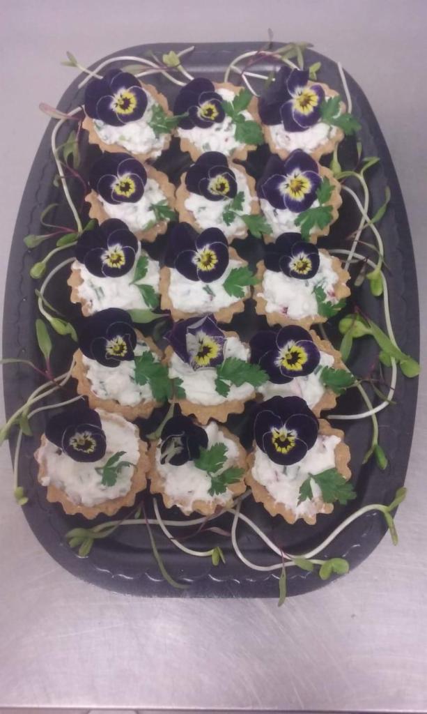 Tarty z kremowym farszem, dekorowane fioletowymi bratkami i zieloną pietruszką, ułożone na czarnej tacy z pędami kiełków.