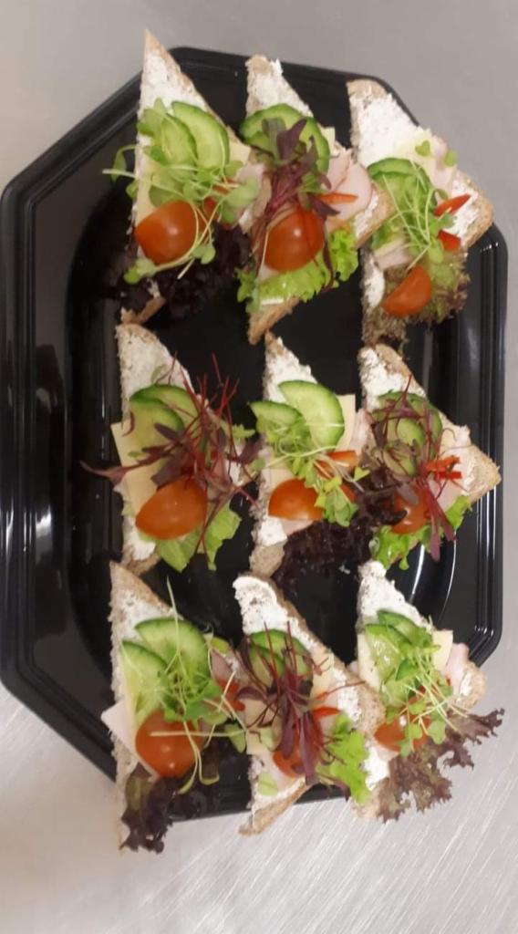 Trójkątne kanapki na czarnym talerzu, z kremowym serem, szynką, serem żółtym, pomidorem koktajlowym, ogórkiem i mikrolistkami. Dekoracyjne ułożenie, widoczne delikatne listki sałaty i kiełki.