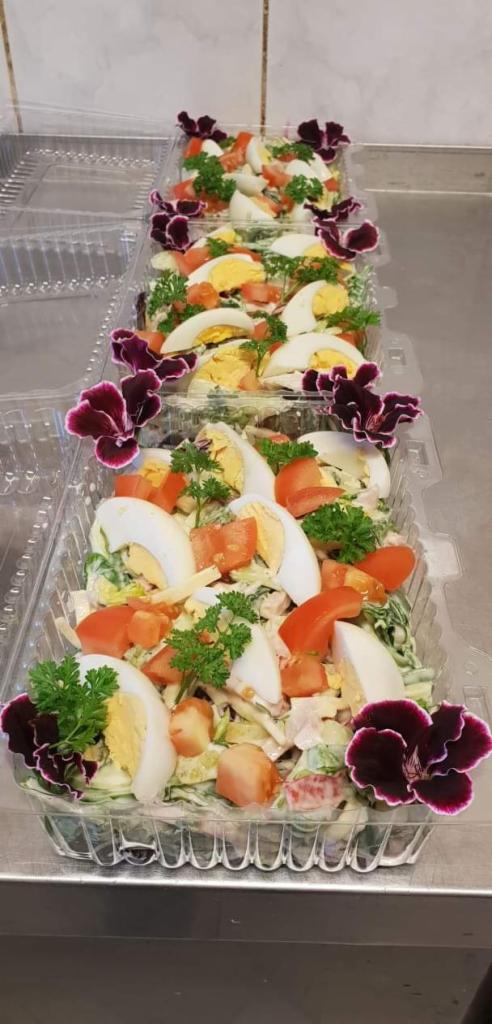 Rząd przezroczystych pojemników z sałatką warzywną, udekorowaną kwiatami, z jajkiem, pomidorami i zieloną pietruszką.