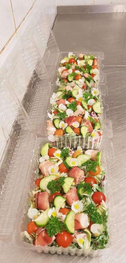 Trzy plastikowe pojemniki z sałatką: szynka parmeńska, pomidory koktajlowe, ogórek, natka pietruszki i białe kwiatki na białym podłożu.