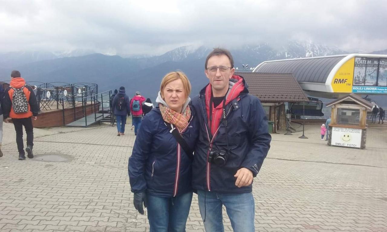 Para na tle kolejki linowej i gór. Mężczyzna z aparatem Sony. Na pierwszym planie bruk, w tle platforma widokowa z barierkami i grupa osób.