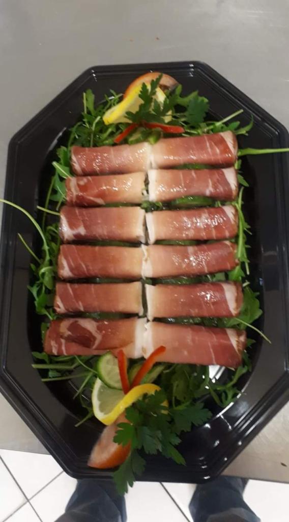 Przystawka z szynki parmeńskiej zawiniętej wokół farszu, ułożona rzędami na czarnym talerzu z rukolą, plastrami cytryny i papryki.