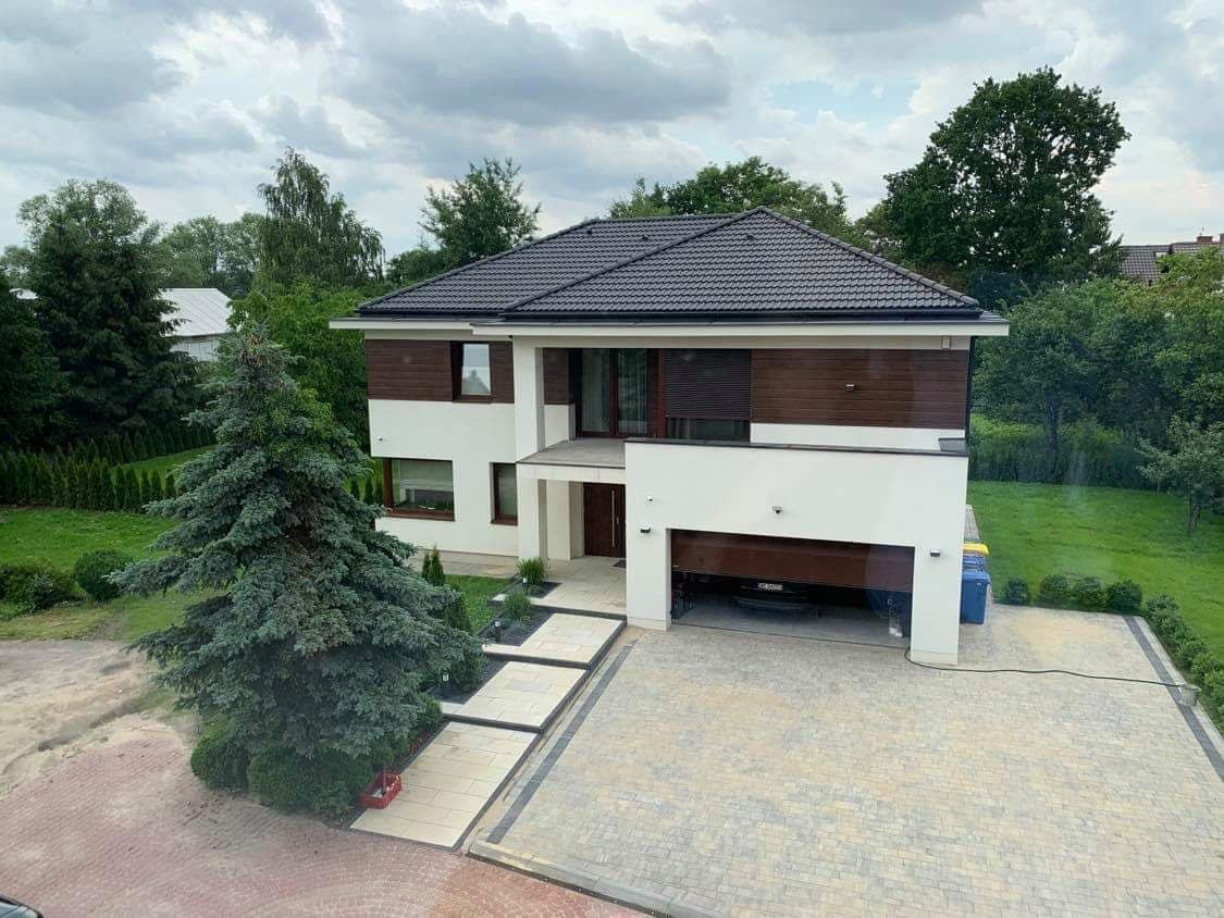 Dwukondygnacyjny dom z płaskim dachem, białymi ścianami i drewnianą okładziną na górnej kondygnacji, z otwartym garażem, podjazdem z kostki brukowej i zielonym trawnikiem z iglakiem z przodu.