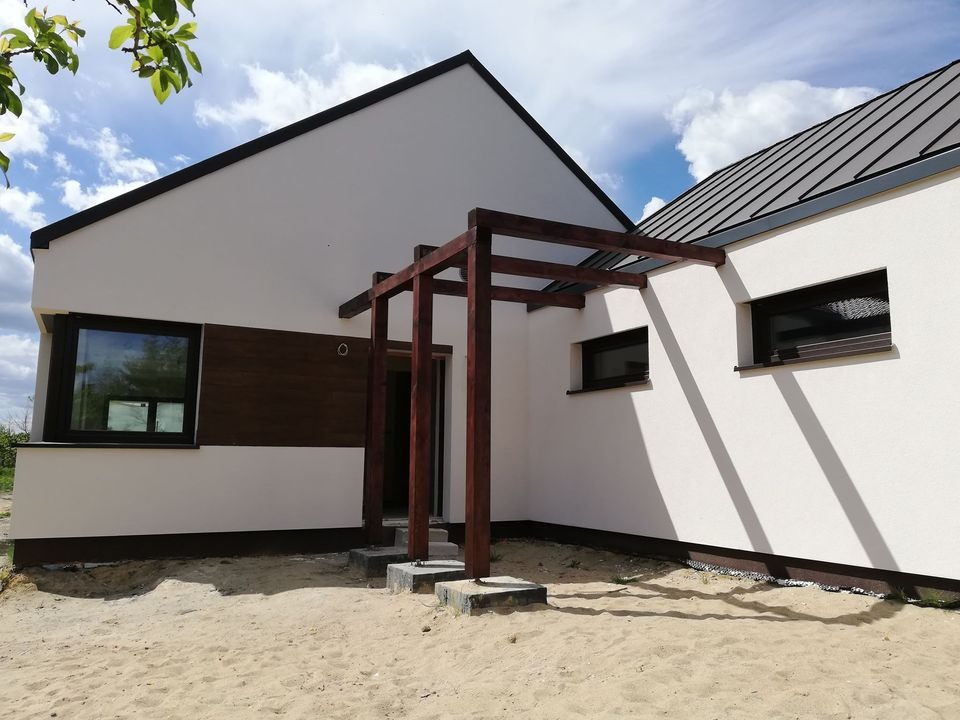 Nowoczesny dom jednorodzinny z kremową elewacją, drewnianą pergolą nad wejściem i elementami z ciemnego drewna, widoczny z zewnątrz na piaszczystym terenie.